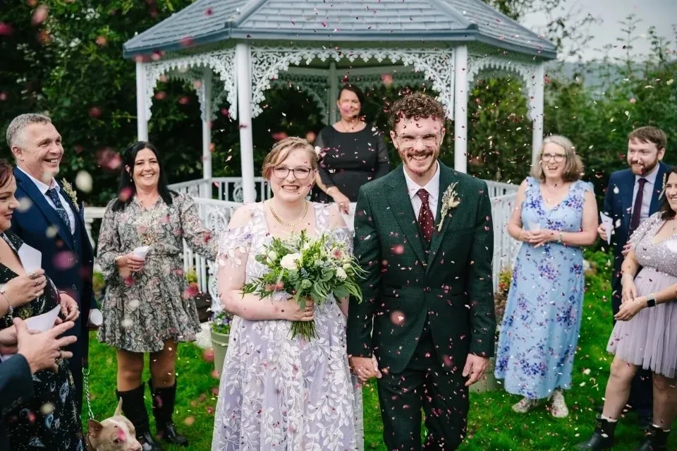 Bride and groom walking through confetti shower in front of the white gazebo surrounded by smiling guests