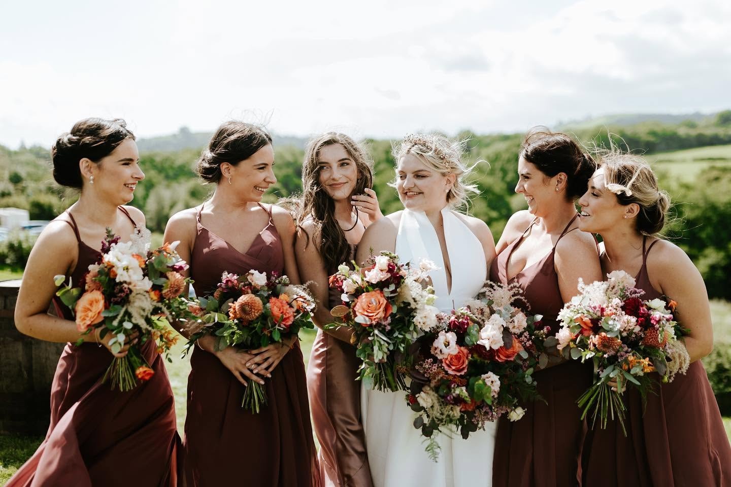 Bride with bridesmaids in dusty rose dresses holding autumn bouquets with Welsh countryside behind them