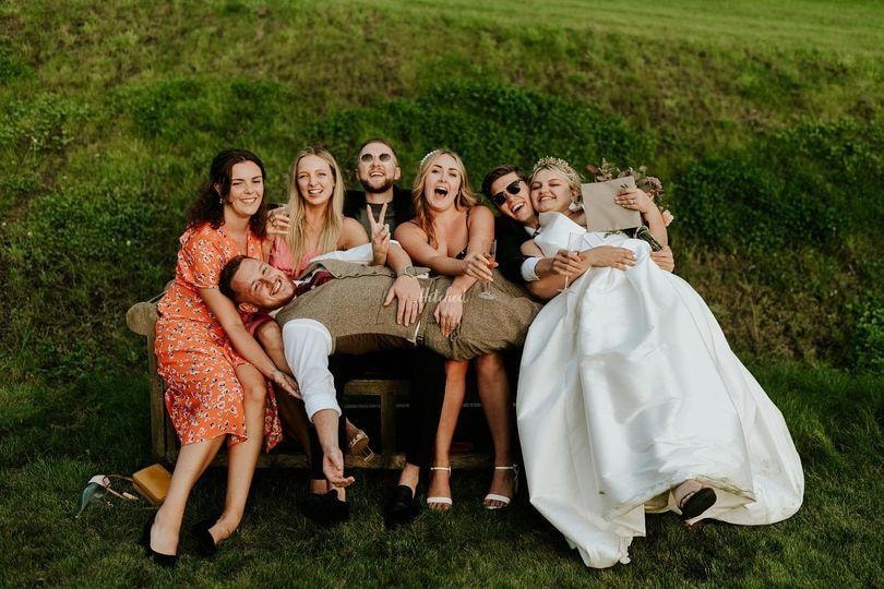 Bride and wedding guests laughing together on a bench in the sunshine at Oak Tree Farm